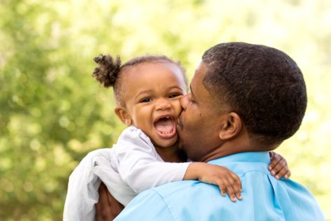 A Black father kisses his daughter in front of a lush green tree. The daughter is smiling wide and wearing a white dress.